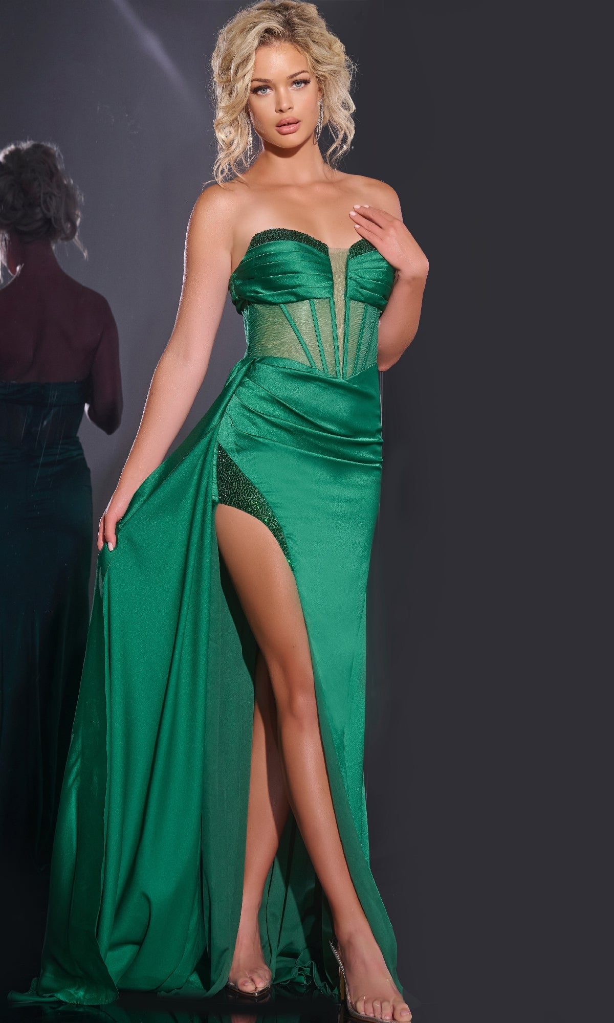 A woman stands on a dark backdrop in the Jovani 43157 Long Prom Dress—a strapless, emerald green gown with a high slit; her reflection is visible behind her.